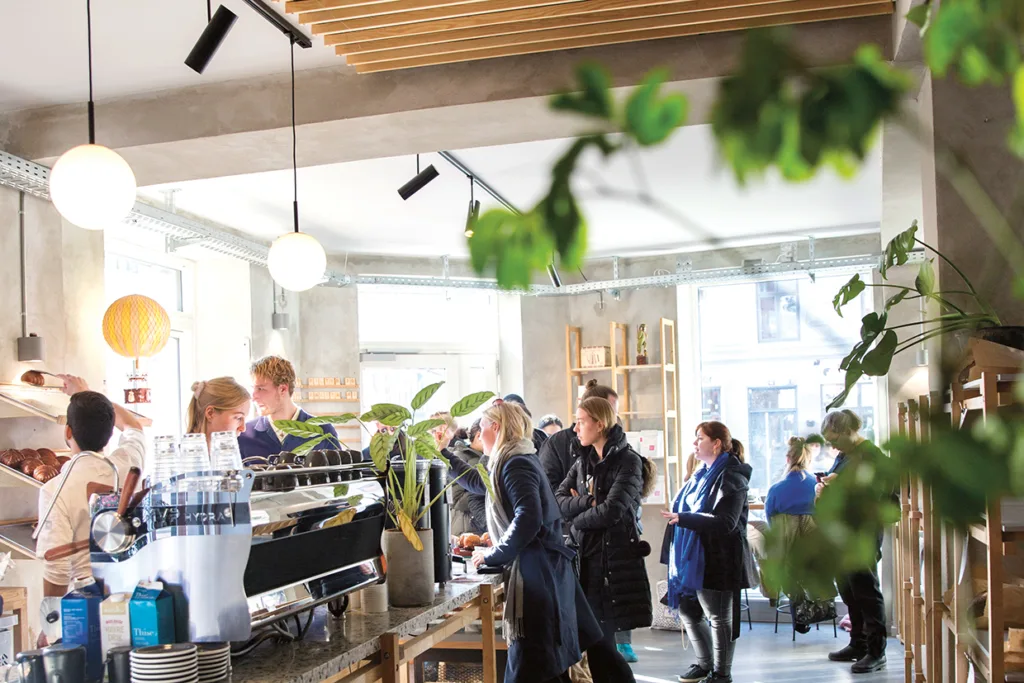 Ett ljust, modernt kafé med människor som står i kö för att beställa vid disken. Solljuset flödar in genom stora fönster och växter och träinredning skapar en mysig atmosfär. Personalen förbereder drycker och bakverk bakom disken.