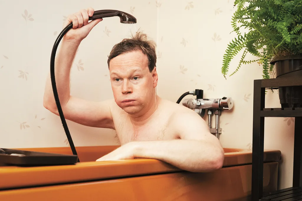 Johan Glans sitter i ett brunt badkar med ett duschhuvud över huvudet och ser förvånad eller frustrerad ut. Bredvid badkaret står en ormbunke i kruka och badrummets väggar har ett ljust blommönster.