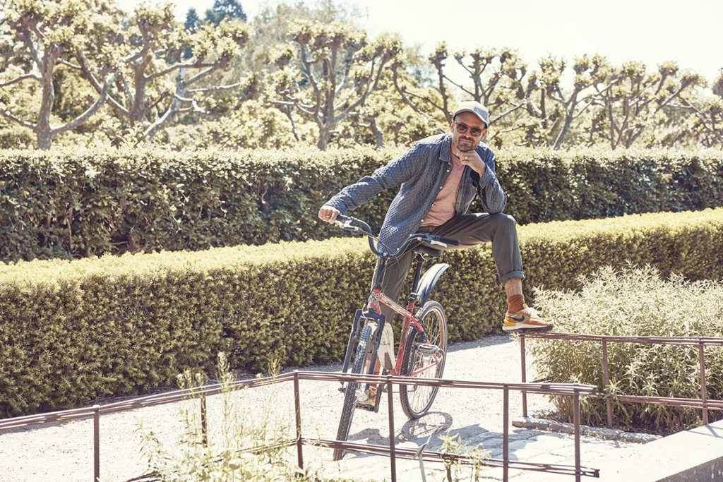 David Dencik i fritidskläder och solglasögon poserar på en cykel, med ena benet på styret, i en solig trädgård med klippta häckar och beskurna träd i bakgrunden.