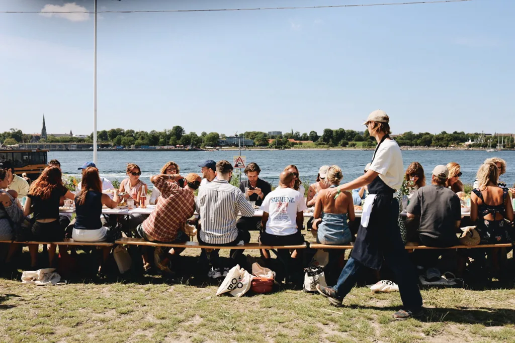 En grupp människor sitter vid ett långt bord utomhus nära vattnet och äter en måltid tillsammans. En servitör med förkläde går förbi och i bakgrunden syns träd och byggnader på andra sidan vattnet.