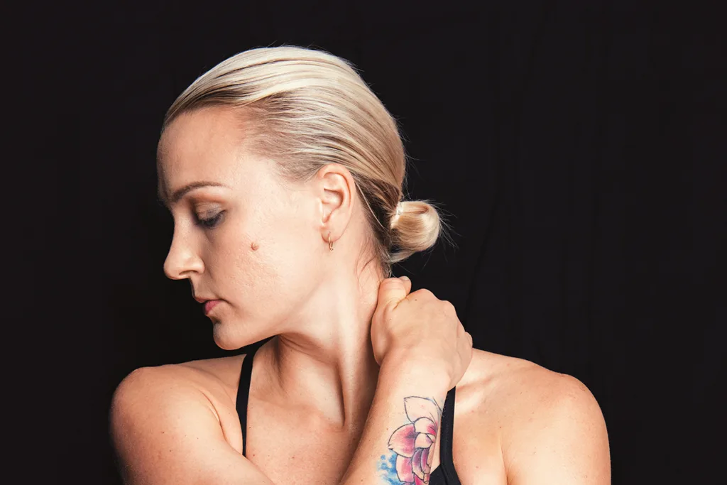 Sara Sjöström med blont hår i en låg knut tittar ner och åt sidan och rör vid sin nacke. Hon har en tatuering med en blomma på underarmen mot en svart bakgrund.