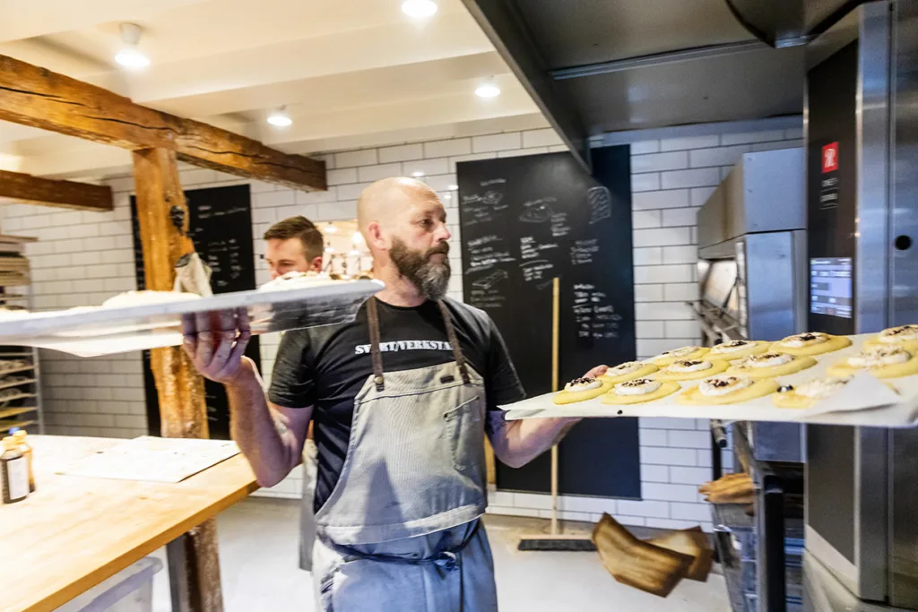 Johan Sörbergi förkläde håller två brickor med obakade bakverk nära en ugn i ett modernt bagerikök, medan en annan person arbetar i bakgrunden.