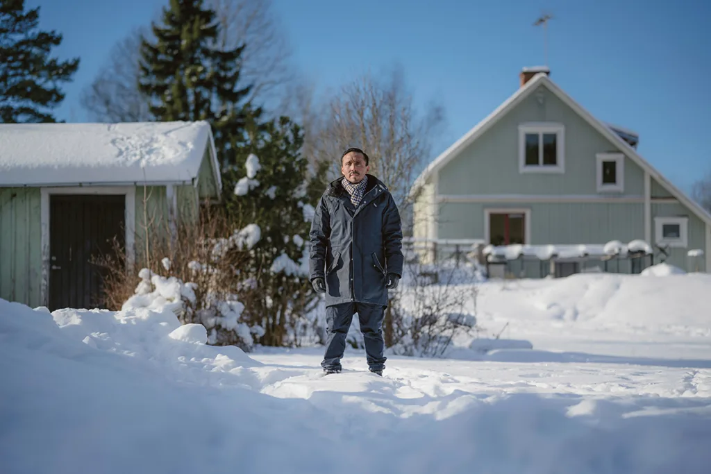 En person i vinterjacka står ute på en snöig gård med två hus och träd i bakgrunden under en klarblå himmel.