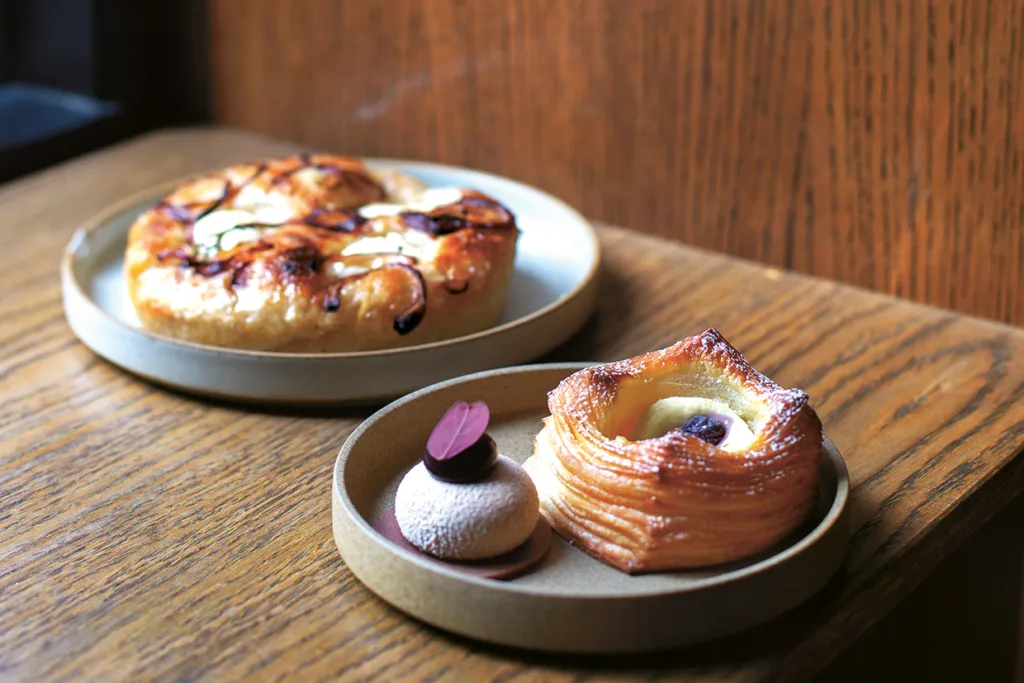 Två tallrikar på ett träbord, den ena med ett glaserat bakverk och den andra med ett fyrkantigt bakverk och en rund dessert som toppas med en chokladskiva.