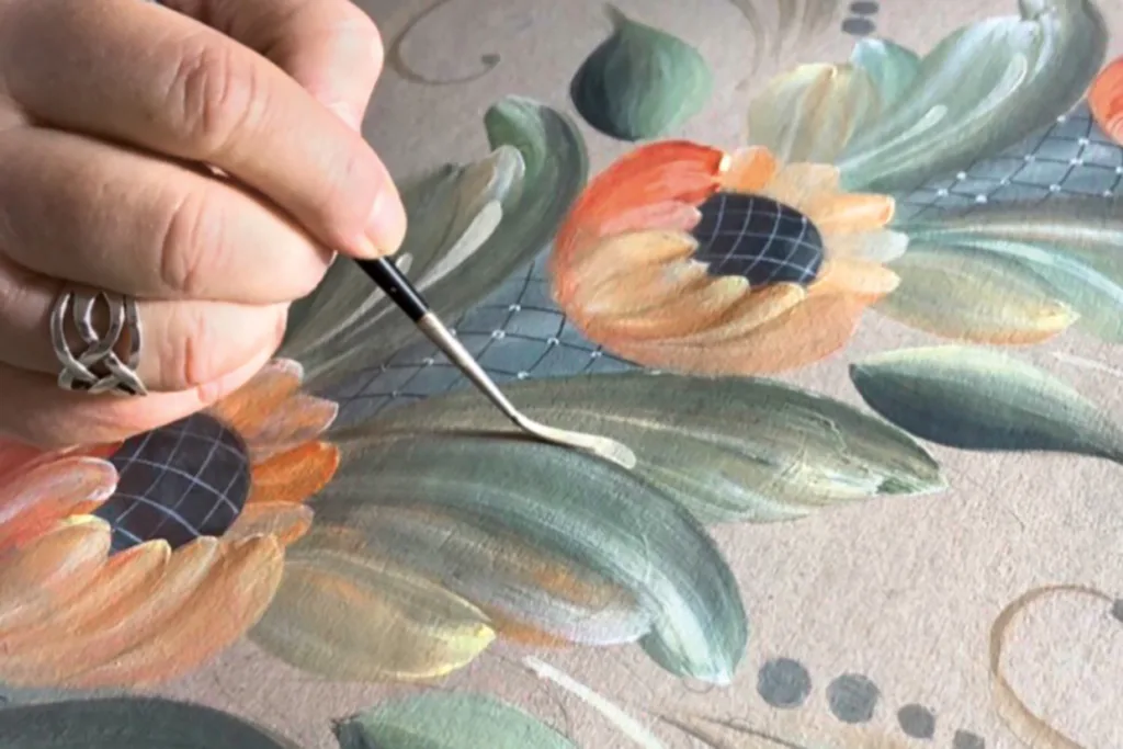 En hand som håller i en pensel lägger till ljusa detaljer på ett grönt blad på en målad blomsterdesign med solrosor och blad på en texturerad yta.