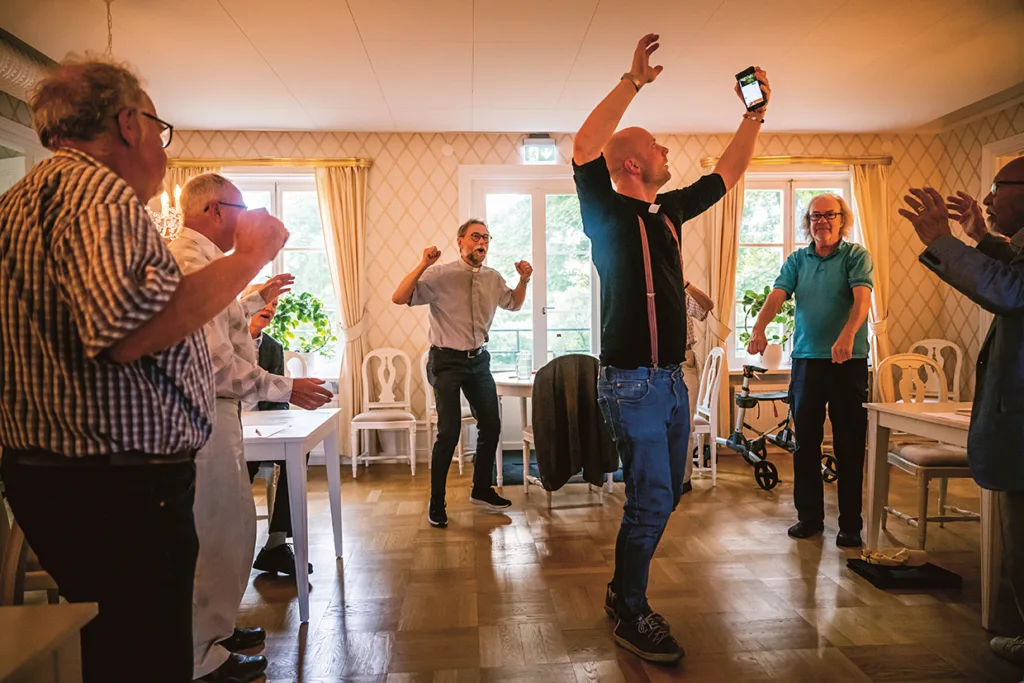 En grupp äldre personer dansar entusiastiskt tillsammans i ett ljust rum med trägolv och stora fönster, medan en man höjer sin telefon och sina armar och leder den livliga aktiviteten.