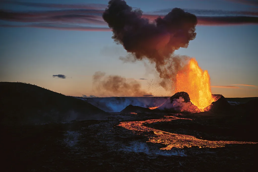 Lava bryter ut och strömmar från en vulkan i skymningen, med en stor plym av mörk rök som stiger upp mot himlen och glödande orange lava som lyser upp landskapet.