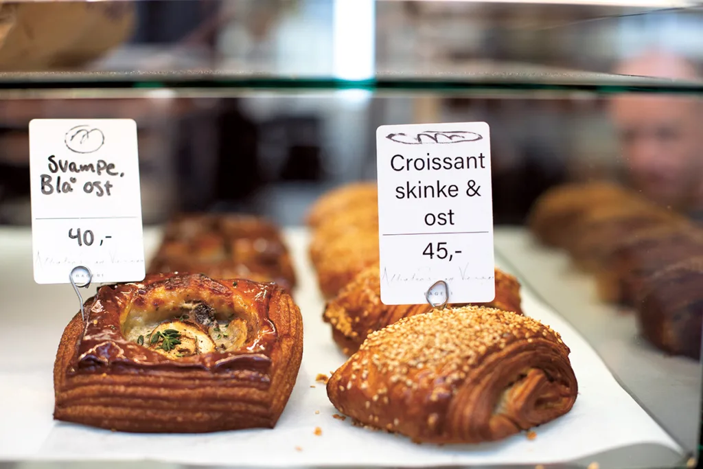 Bagerimonter med bakverk märkta Svampe, Blå ost och Croissant skinke & ost på skyltar. Bakverken är gyllenbruna, vissa med frön och synliga fyllningar. Priserna är angivna i danska kronor.