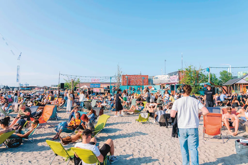 En stor skara människor kopplar av på färgglada stolar och bänkar på en sandstrand under en klarblå himmel, med containrar och ljusslingor i bakgrunden.