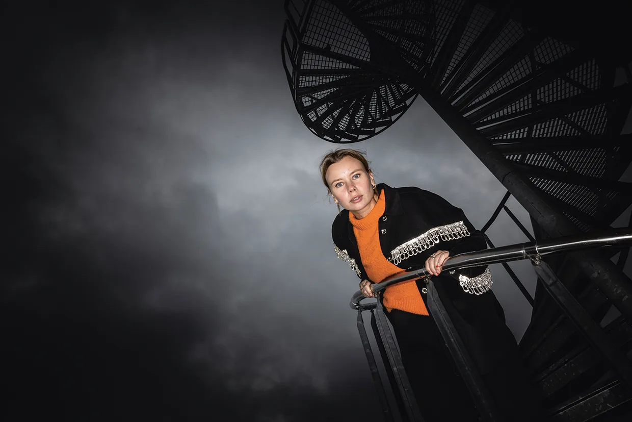 Amanda Jansson i orange tröja och mörk rock med dekorativa kanter lutar sig mot ett räcke och tittar ner, med en spiraltrappa och en dramatisk, molnig himmel i bakgrunden.