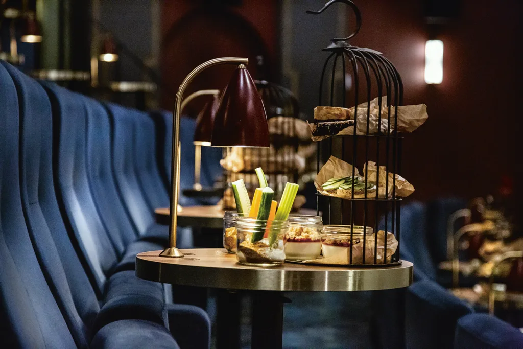Ett runt bord i en svagt upplyst lounge med stolar i blå sammet, en lampa, glasburkar med selleristavar och dipp och en fågelbur i två våningar med smörgåsar på brunt papper.