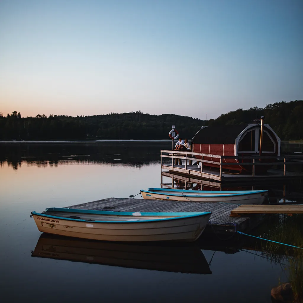 Två roddbåtar ligger förtöjda vid en träbrygga på en lugn sjö i solnedgången, med en liten röd byggnad och solstolar på bryggan, och träd i bakgrunden.