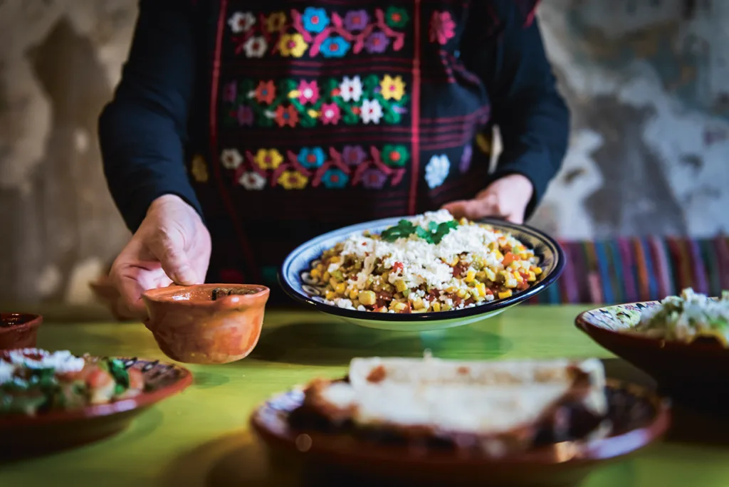 En person i en färgglad broderad topp serverar en tallrik med mexikansk mat med majs, bönor och ost, tillsammans med en liten lerskål, på ett grönt bord med fler maträtter.