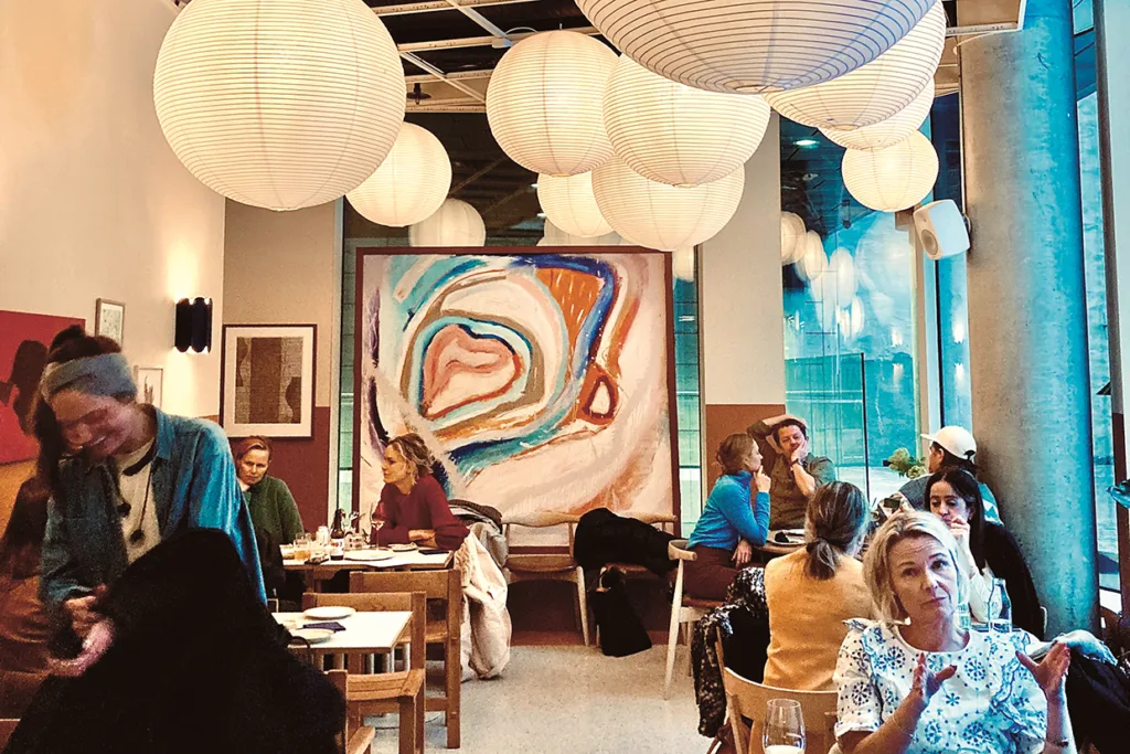 Människor äter middag på en modern restaurang med runda papperslyktor hängande från taket och abstrakta konstverk på väggen. Naturligt ljus flödar in genom höga fönster och skapar en livlig och mysig atmosfär.