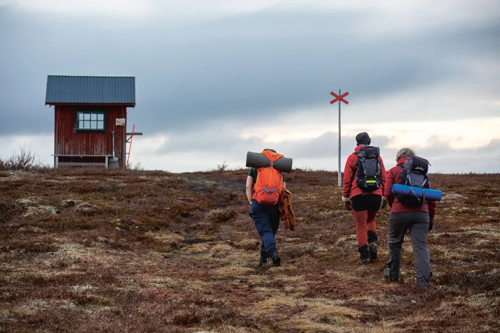 Tre vandrare med ryggsäckar och utrustning går mot en liten röd stuga på ett gräsbevuxet, stenigt landskap under en molnig himmel, med en röd ledmarkering synlig framför dem.