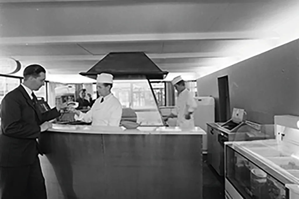 En man i kostym beställer mat från en arbetare i vit uniform och hatt bakom en cafeteriadisk, medan en annan arbetare ses i bakgrunden nära köksutrustning.