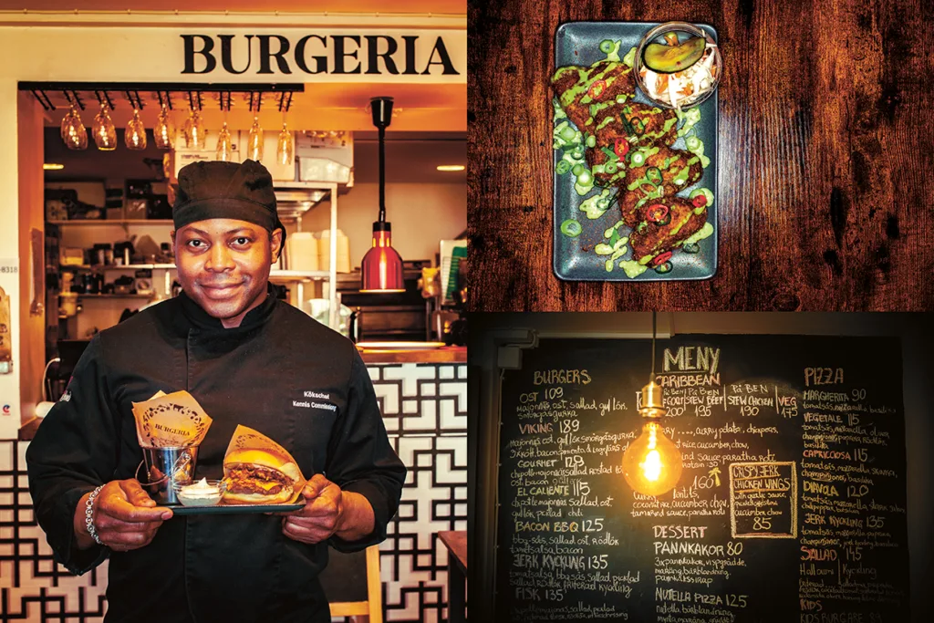 En kock i svart uniform håller i två hamburgare vid en restaurangdisk märkt BURGERIA. Bredvid honom syns en tallrik med mat och en meny med hamburgare och pizza på en griffeltavla under en hängande glödlampa.