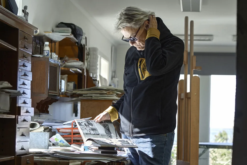 Lars Lerin, iförd solglasögon, mörk jacka och jeans står i ett rörigt rum och tittar på en uppslagen bok på ett skrivbord, med ena handen vilande på boken och den andra vidrörande nacken.