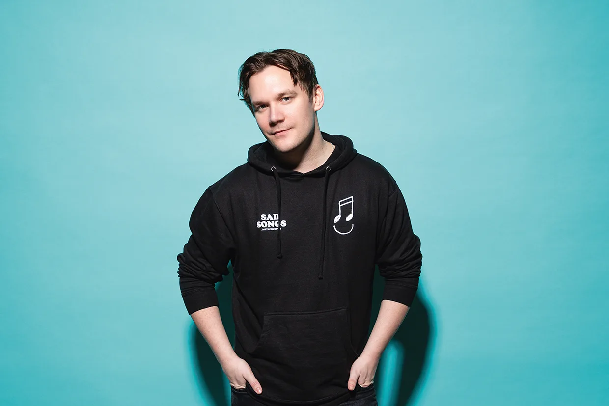 Christopher Garplind med kort brunt hår står mot en ljusblå bakgrund, klädd i en svart huvtröja med vita mönster, inklusive orden SAD SONGS och en musiknot, med händerna i fickorna och ett litet leende.
