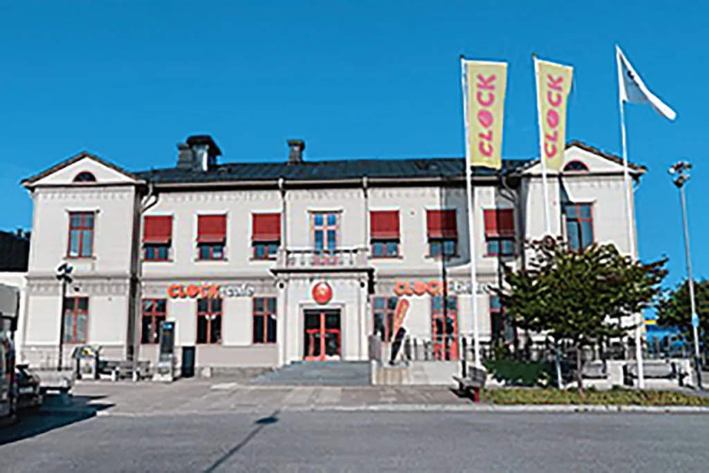 En tvåvåningsbyggnad med röda fönsterluckor och tre flaggor med texten CLOCK på framsidan. Fasaden har CLOCK-logotyper ovanför dörrarna och fönstren, och det finns ett träd och några parkerade bilar i närheten under en klarblå himmel.