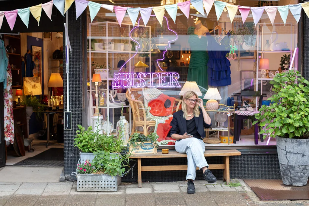 En kvinna sitter på en bänk utanför en vintagebutik som är dekorerad med färgglada banderoller och växter, med en neonskylt och olika kläder och lampor synliga genom fönstret bakom henne.