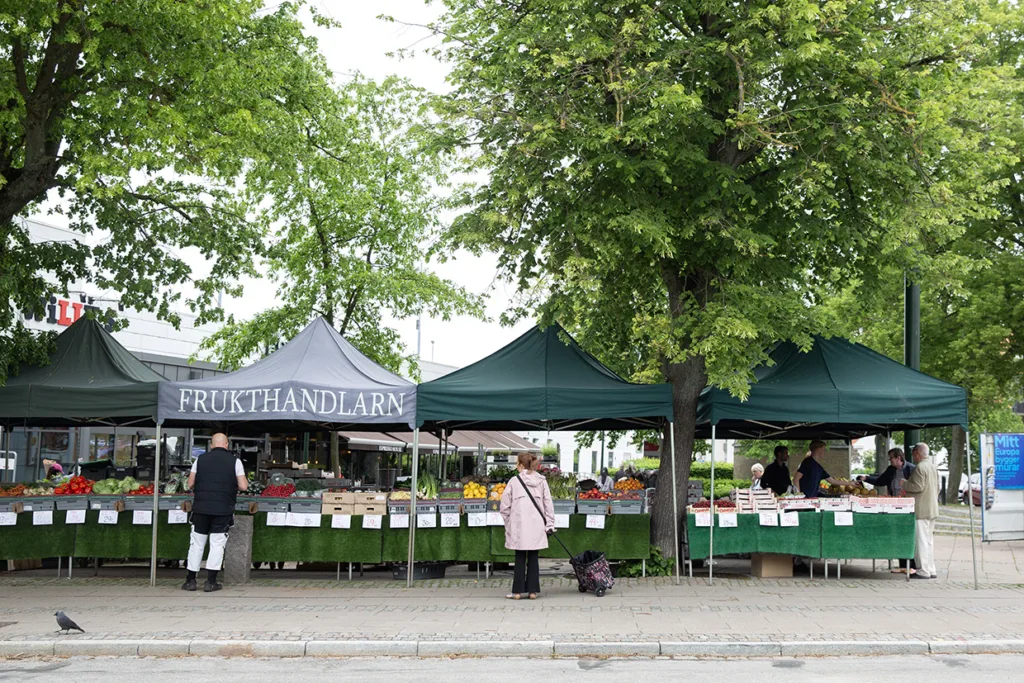 Utomhusmarknad med gröna tält märkt Frukthandlarn. Flera personer tittar på bord fyllda med frukt och grönsaker. Träd kantar trottoaren och en duva promenerar på trottoaren i förgrunden.