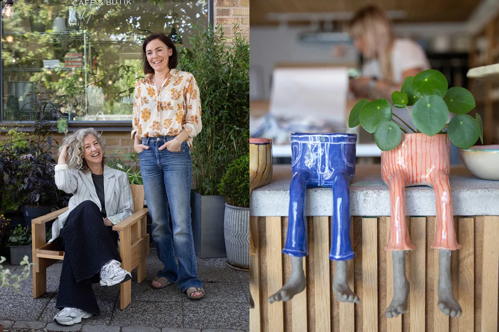 Två kvinnor ler utanför ett café, den ena sittande och den andra stående. Bredvid dem står udda blomkrukor i form av färgglada byxor med fötter på en trädisk, med en suddig person i bakgrunden.