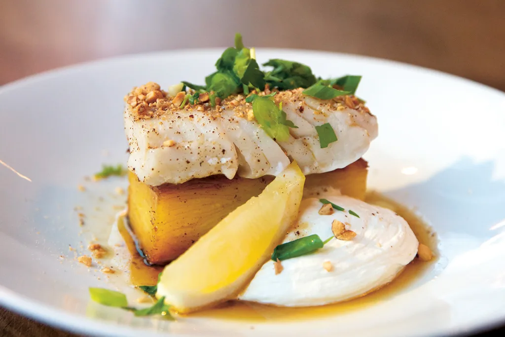 En tallriksrätt med en bit vit fisk på en gyllene rostad potatis, garnerad med hackade gröna örter och krossade nötter, serverad med en klick krämig sås och en citronklyfta.