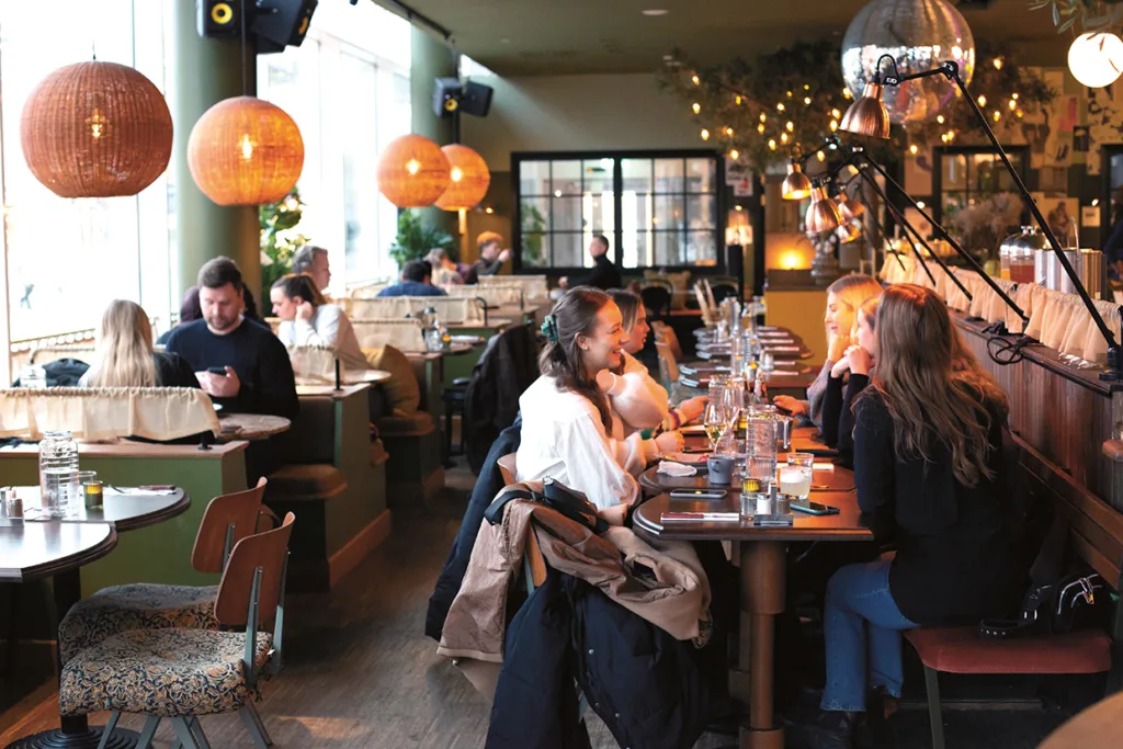 En mysig restaurang med varm belysning och hängande lampor, där grupper av människor sitter vid bord och äter, dricker och pratar i en avslappnad atmosfär.