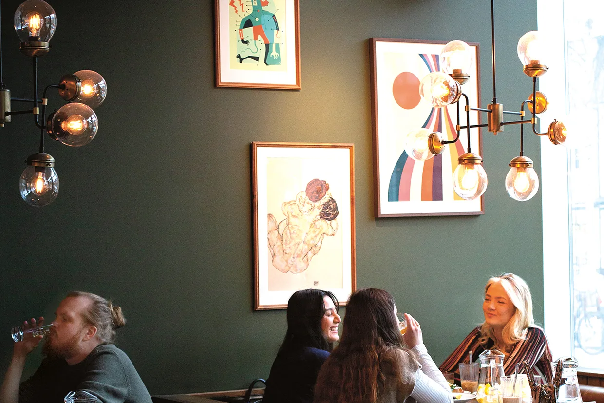 Tre kvinnor sitter och pratar vid ett restaurangbord med drinkar och ler, medan en man sitter bredvid och dricker vatten. Modern konst och hängande lampor pryder den gröna väggen bakom dem.