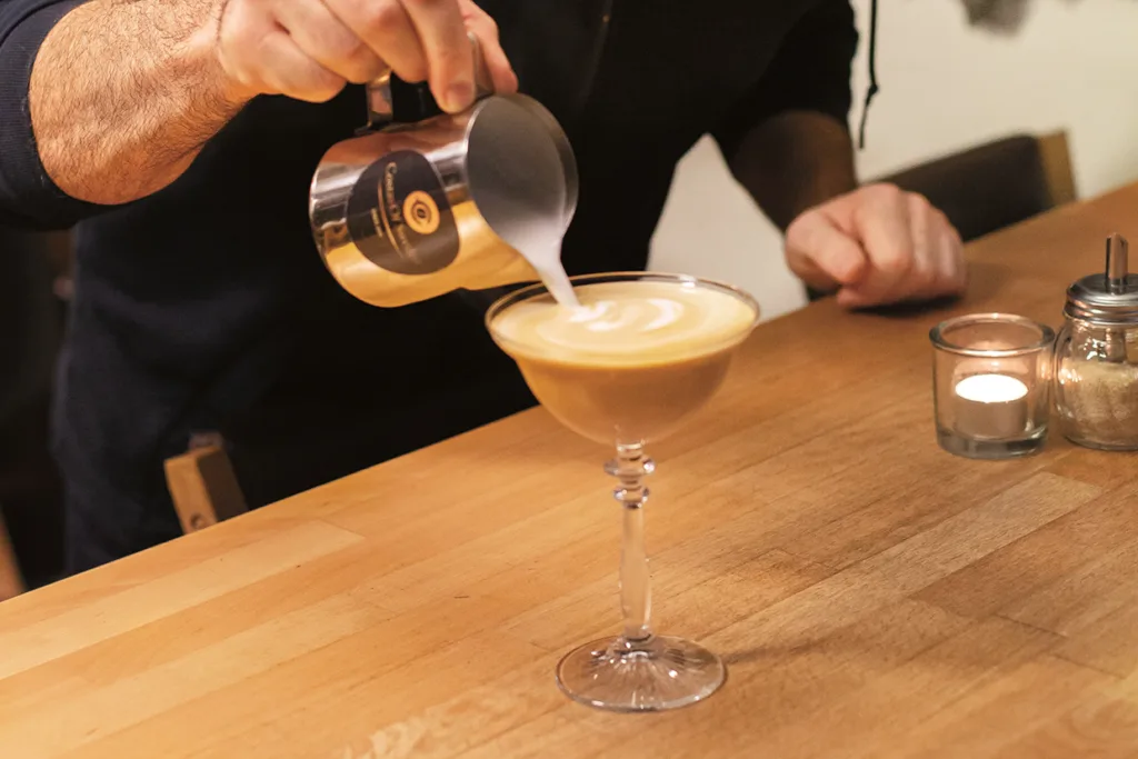En person häller upp ångad mjölk från en metallkanna i ett coupeglas med kaffe på en träbänk bredvid ett tänt ljus och en sockerburk.