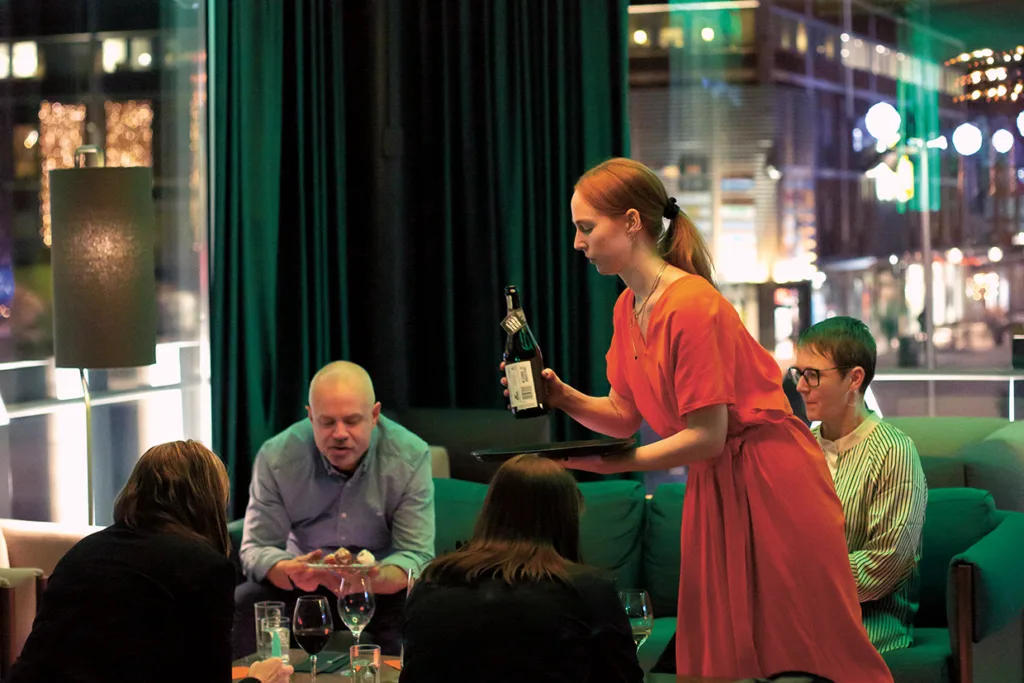 En servitris i röd klänning presenterar en flaska vin till ett bord med fyra personer som sitter i en modern, svagt upplyst restaurang med stora fönster och stadsljus som syns utanför.