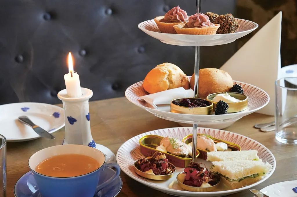 På en tredelad bricka ligger olika bakverk, scones och smörgåsar på ett träbord dukat för afternoon tea, med ett tänt ljus och en kopp te bredvid.