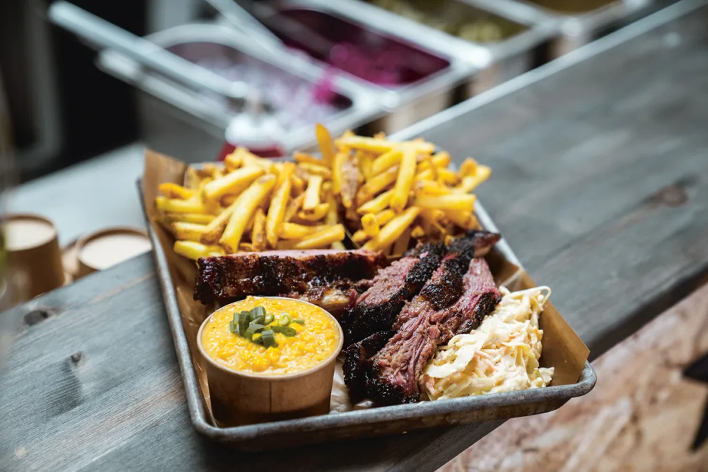 En bricka med grillmat med skivad brisket, revbensspjäll, gyllene pommes frites, en portion krämig makaroner och ost toppad med salladslök och en portion coleslaw på ett rustikt träbord.