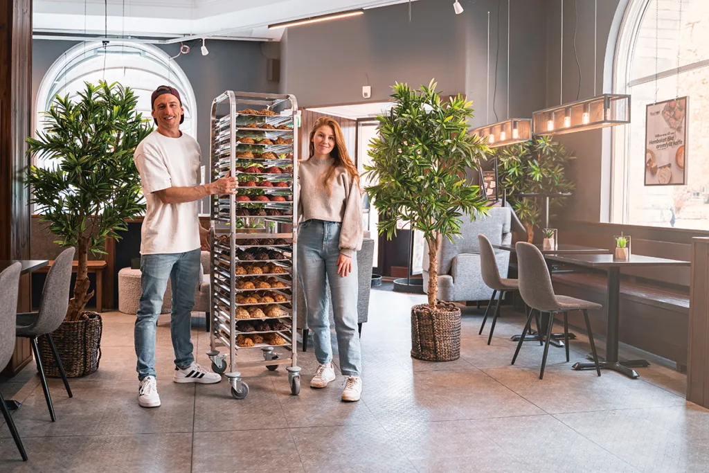 Två personer står leende på ett modernt kafé och håller i ett högt ställ fyllt med brickor med bakverk. Kaféet har stora fönster, grå väggar och flera krukväxter som dekorerar utrymmet.