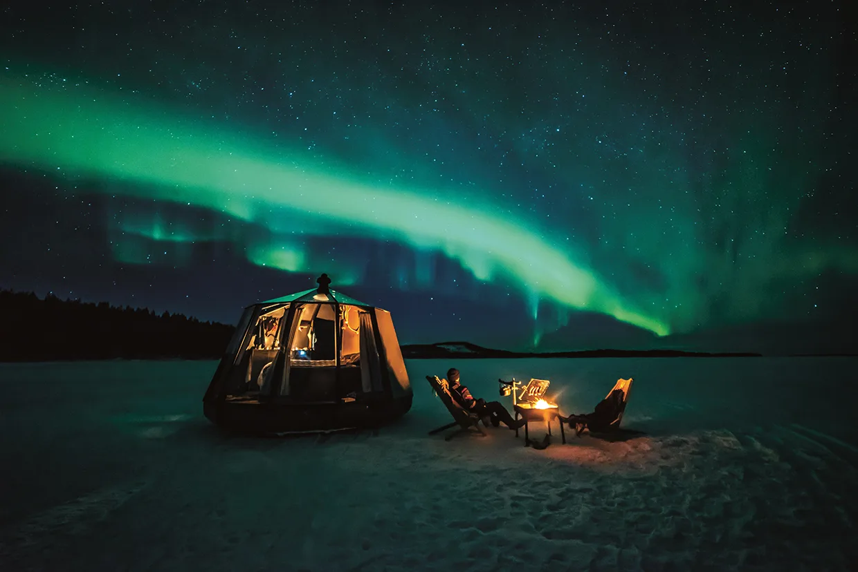 Två personer kopplar av vid en brasa på snöig mark bredvid ett mysigt tält och ser gröna norrsken dansa över en stjärnfylld natthimmel.