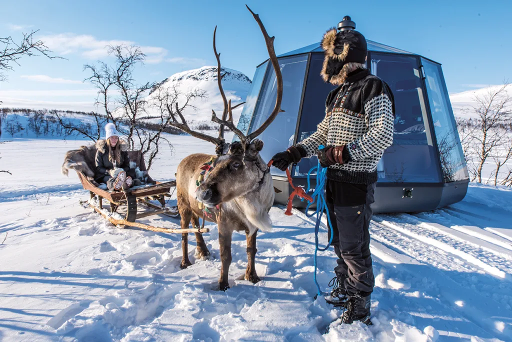 En person i vinterkläder står och håller i renens tyglar, medan en annan person och ett barn sitter på en träsläde i ett snölandskap med berg och en modern glashytta i bakgrunden.