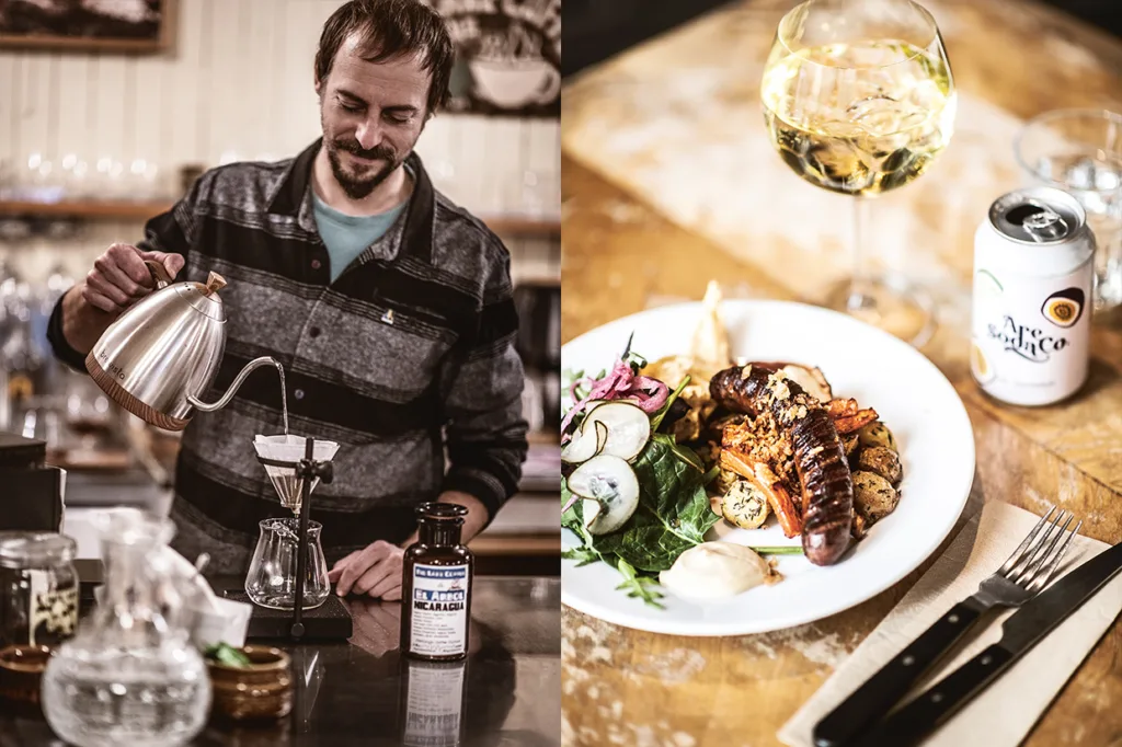 En man häller upp varmt vatten i en kaffedropp vid en disk. Bredvid står en tallrik med grillad korv med grönsaker, ett glas vitt vin och en burk, med bestick på en servett bredvid.