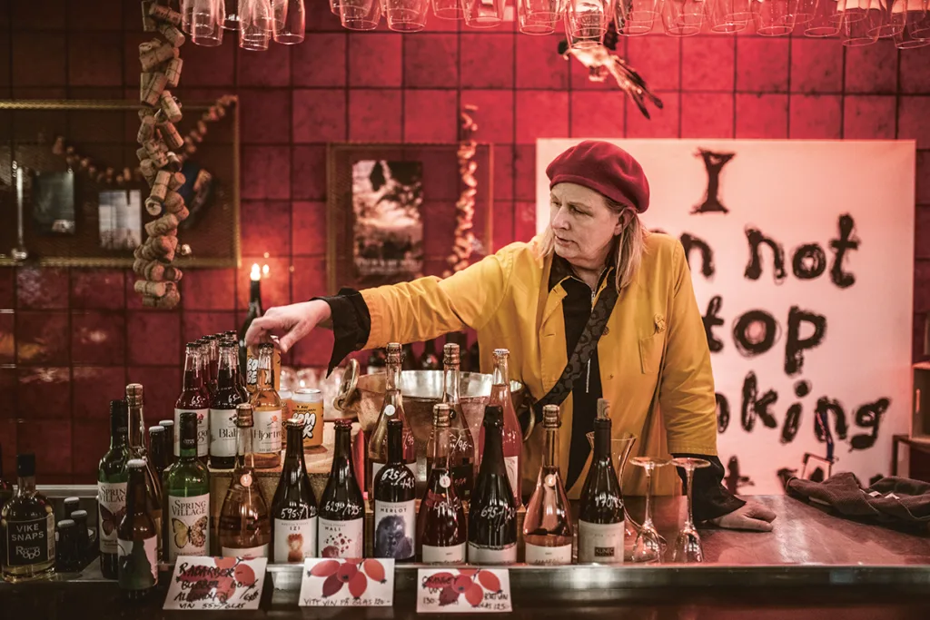 En person i gul jacka och röd basker står bakom en bar med olika flaskor. Baren är svagt upplyst och dekorerad med korkar, ljus och en stor skylt med texten "Jag kan inte sluta dricka".