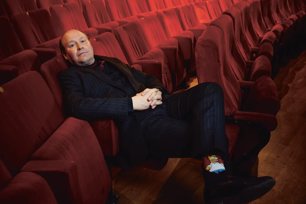 En skallig Henrik Dorsin i mörk kostym sitter avslappnad i en tom biosalong och lutar sig tillbaka med händerna i kors. De röda sätena omger honom och han bär färgglada strumpor med en seriefigur.