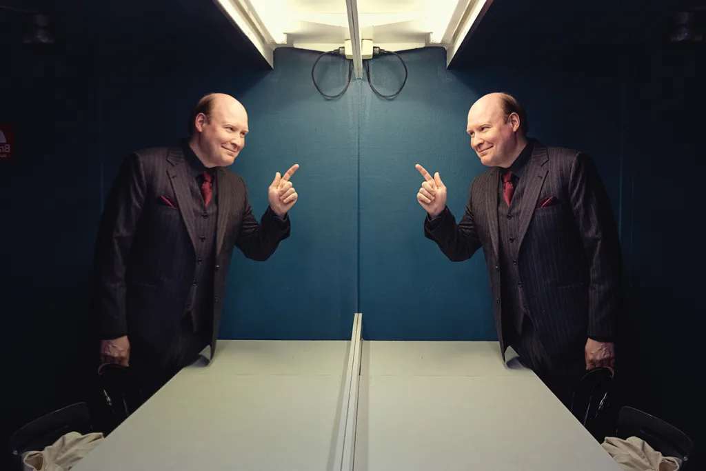 En kostymklädd Henrik Dorsin står framför en spegel, ler och pekar på sin egen spegelbild med båda händerna. Rummet har blå väggar och är upplyst av fluorescerande lampor ovanför.