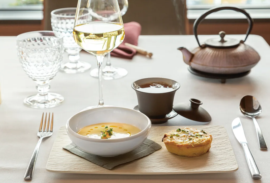 På en träskiva på ett dukat matbord finns en skål med krämig soppa, en liten quiche, ett glas vitt vin, en kopp te med tekanna och bestick, vilket skapar en elegant måltidspresentation.