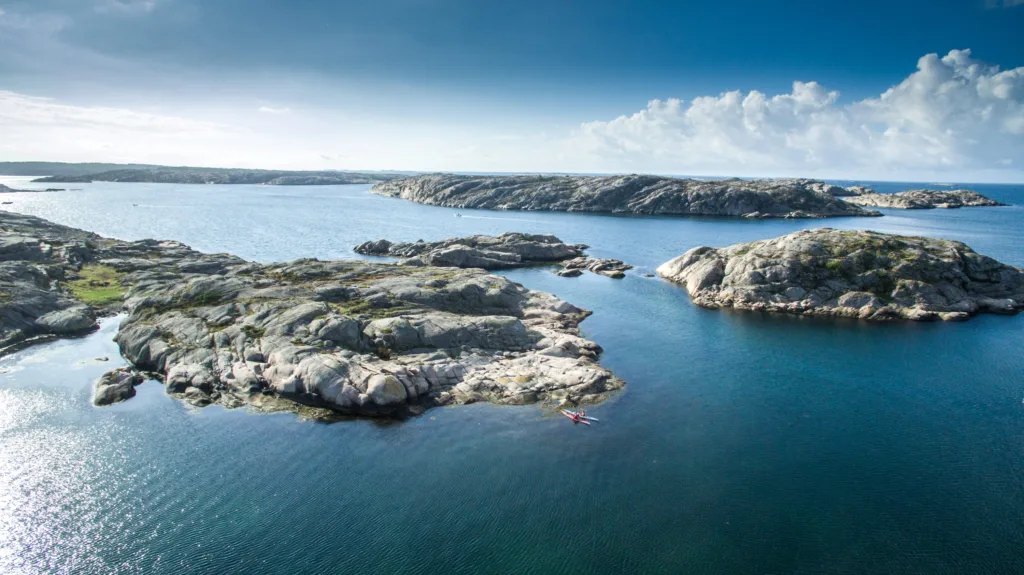 Flygfoto över ett lugnt blått hav med klippiga öar utspridda över hela havet. En person i en röd kajak paddlar nära klipporna under en ljus himmel med spridda moln.
