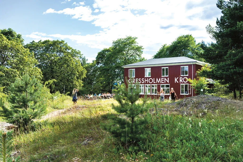 En röd byggnad märkt "GRESSHOLMEN KRO" ligger bland gröna träd och gräsbevuxna fält under en delvis molnig himmel. Människor har samlats vid utomhusbord nära byggnaden och en person går på en stig i närheten.