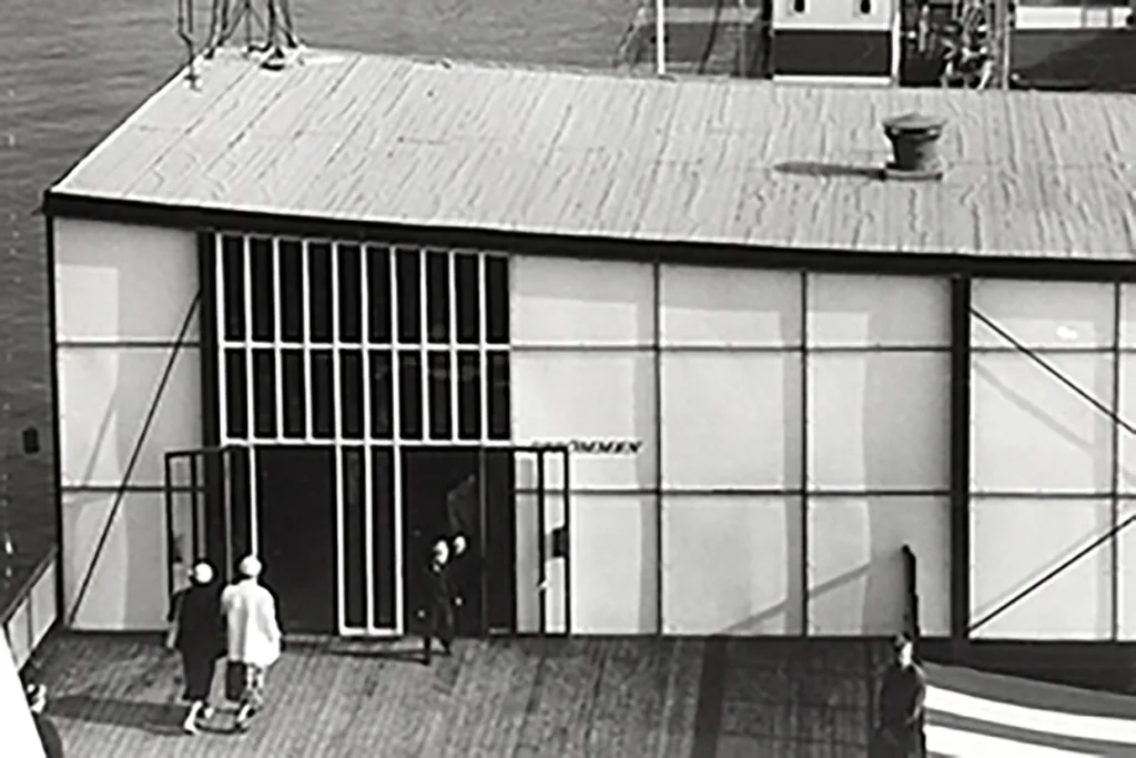 Ett svartvitt foto visar tre personer som går mot entrén till en modernistisk byggnad med stora fönster och ett platt tak, placerad på ett trädäck vid vattnet.