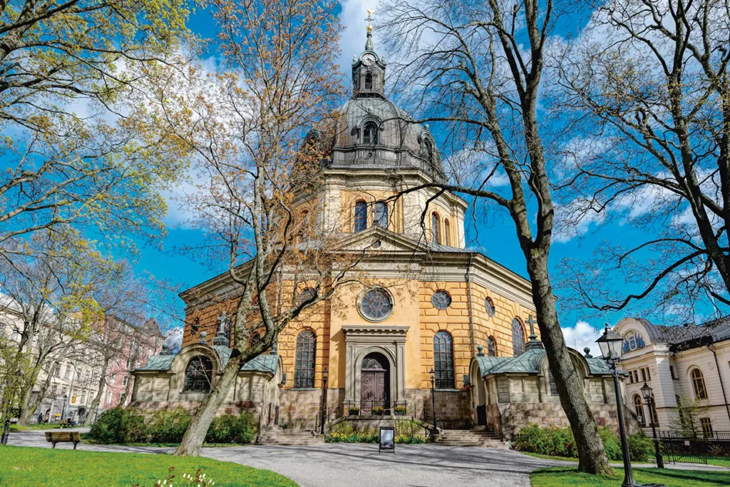 En stor, historisk kyrka med kupoltak står omgiven av träd och en grön gräsmatta under en ljusblå himmel med moln. Byggnaden har välvda fönster, utsmyckade detaljer och gångar som leder fram till entrén.