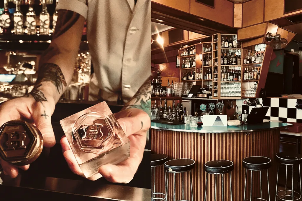 En bartender med tatuerade armar håller en stor, klar isbit och en stämpel vid en bar; bredvid finns ett tomt barområde i retrostil med stolar, flaskor och glas på hyllor.