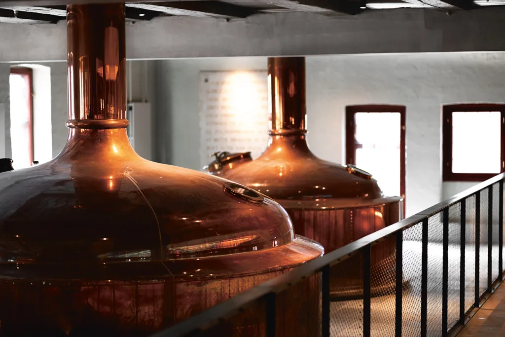 Stora bryggkittlar i koppar inne i ett bryggeri, placerade intill en gångväg med räcken och upplysta av naturligt ljus från närliggande fönster.
