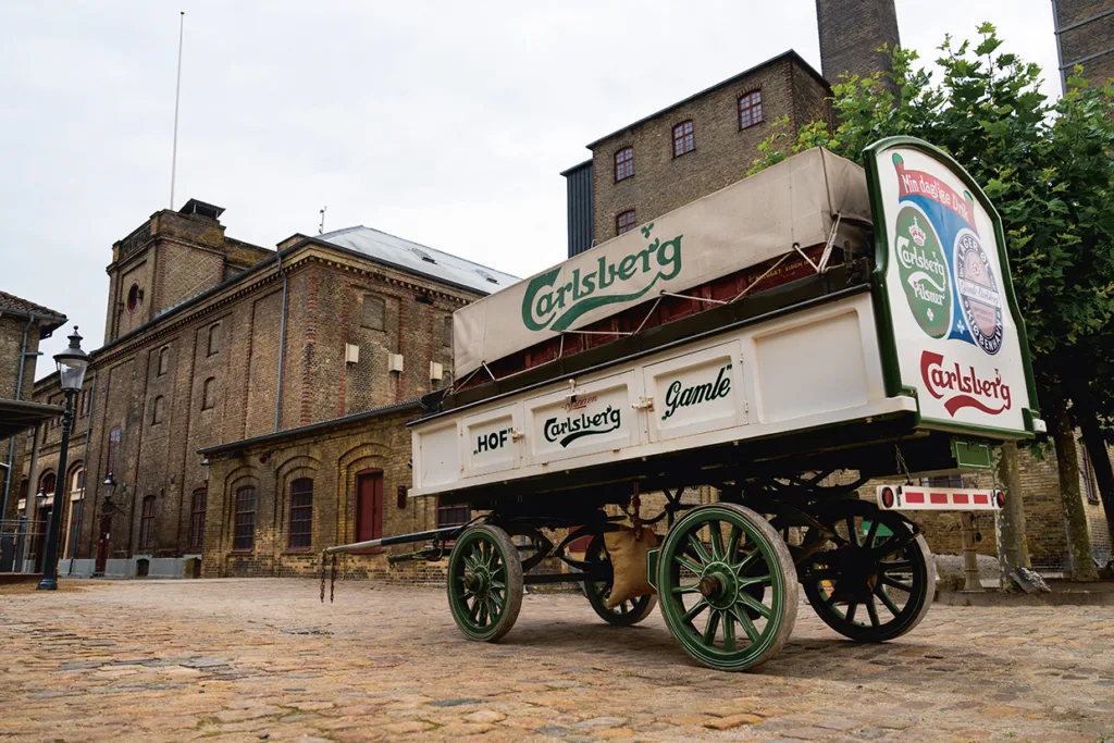 En gammaldags Carlsberg-ölvagn med gröna hjul står på en kullerstensgata framför historiska bryggeribyggnader i tegel. Vagnen är försedd med Carlsberg-logotyper och dansk text.