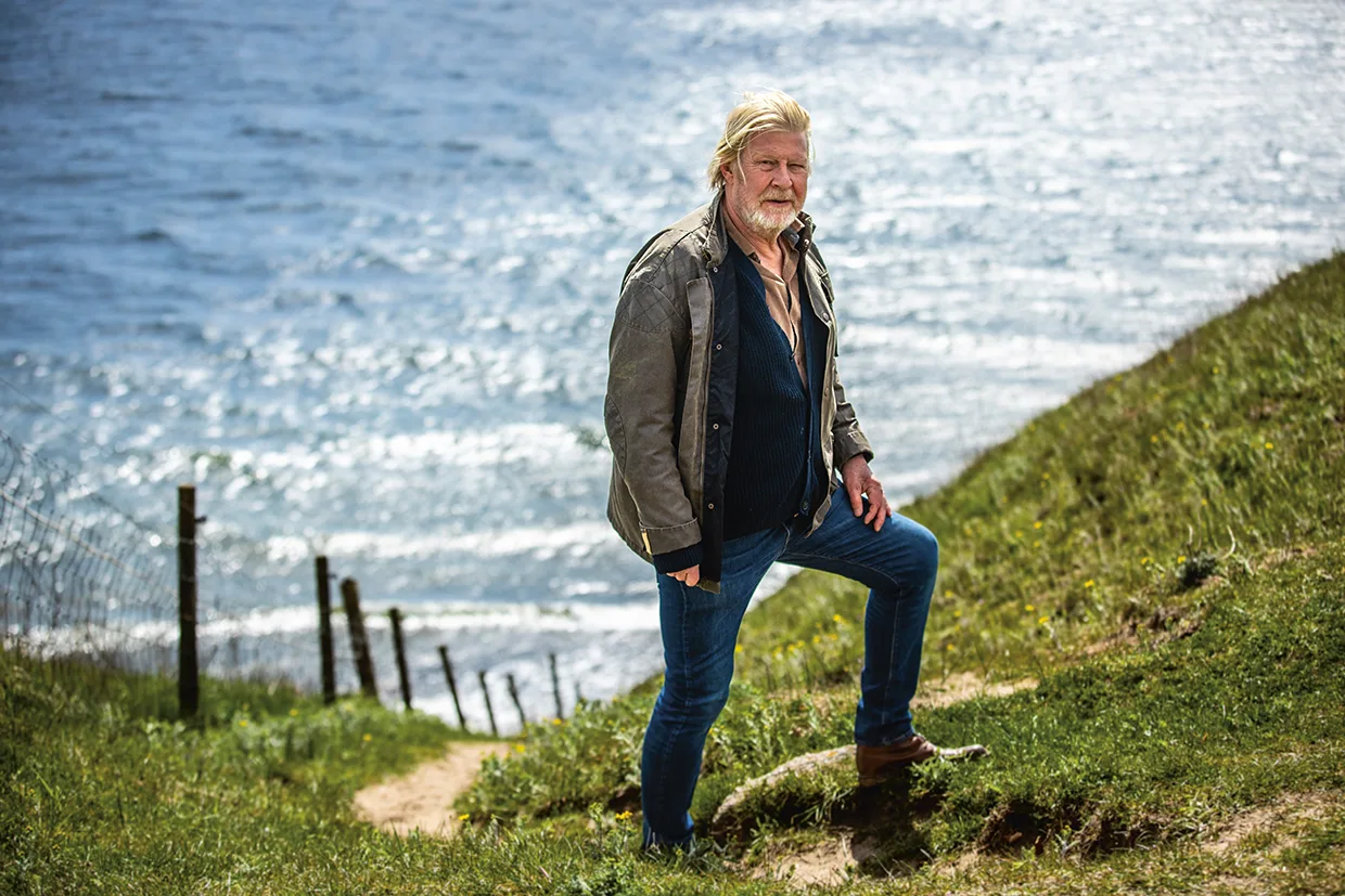 Rolf Lassgård med långt blont hår och skägg står på en gräsbevuxen kulle med utsikt över havet, iklädd jeans, mörk skjorta, jacka och stövlar. Havet och himlen är ljusa i bakgrunden.